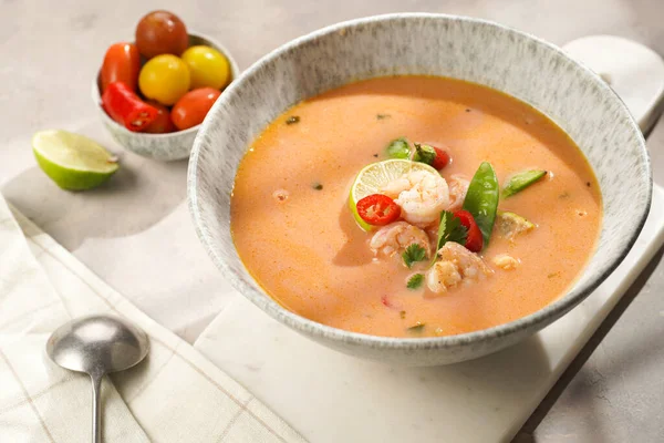 a spicy thai coconut soup or curry made of coconut milk, cherry tomatoes, snow pea, chili pepper, lime, ginger, shrimps and fresh cilantro on marble board on grey concrete background