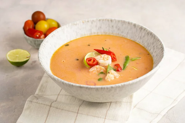 a spicy thai coconut soup or curry made of coconut milk, cherry tomatoes, snow pea, chili pepper, lime, ginger, shrimps and fresh cilantro on kitchen towel on grey concrete background