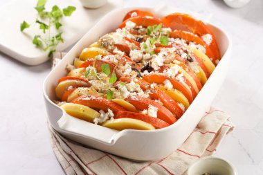 Dilimlenmiş, katmanlı patates ve balkabağı güveci mermer arka planda beyaz bir fırın şeklinde, çiğ malzemeler, üzerine kızarmış soğan, peynir kırıntıları ve krem şanti, fırına hazır.