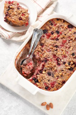 breakfast casserole idea - oven berry baked oatmeal with almonds, walnuts and coconut flakes in a white porcelain baking form on a marble board, two spoons, top view