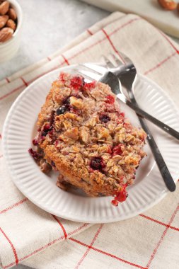 breakfast casserole idea - a piece of oven berry baked oatmeal with almonds, walnuts and coconut flakes on a white porcelain dessert plate on a checkered kitchen napkin, top view