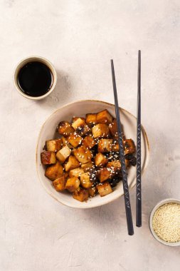 Sticky tofu - vegan soybean protein source in soy sauce and sesame oil marinade roasted on a pan sprinkled with sesame seeds, in a beige colored bowl on a concrete background, top view