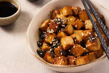Sticky tofu - vegan soybean protein source in soy sauce and sesame oil marinade roasted on a pan sprinkled with sesame seeds, in a beige colored bowl on a concrete background