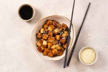 Sticky tofu - vegan soybean protein source in soy sauce and sesame oil marinade roasted on a pan sprinkled with sesame seeds, in a beige colored bowl on a concrete background, top view