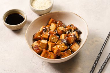Sticky tofu - vegan soybean protein source in soy sauce and sesame oil marinade roasted on a pan sprinkled with sesame seeds, in a beige colored bowl on a concrete background
