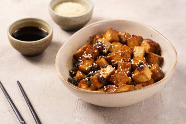 Sticky tofu - vegan soybean protein source in soy sauce and sesame oil marinade roasted on a pan sprinkled with sesame seeds, in a beige colored bowl on a concrete background
