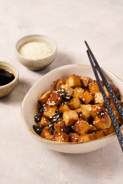 Sticky tofu - vegan soybean protein source in soy sauce and sesame oil marinade roasted on a pan sprinkled with sesame seeds, in a beige colored bowl on a concrete background