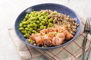 A blue bowl with quinoa tricolore, roughly chopped and pulled salmon and steamed green soy beans edamame on light background