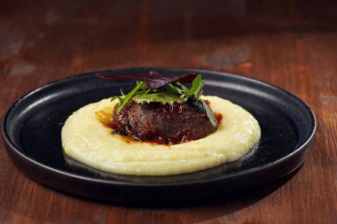 A medium rare ostrich steak in barbecue sauce on a cloud of herb mashed potato topped with fresh green arugula leaves on a black plate on a wooden background