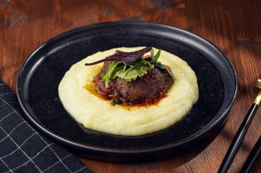 A medium rare ostrich steak in barbecue sauce on a cloud of herb mashed potato topped with fresh green arugula leaves on a black plate on a wooden background