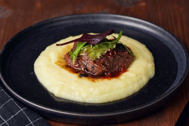 A medium rare ostrich steak in barbecue sauce on a cloud of herb mashed potato topped with fresh green arugula leaves on a black plate on a wooden background