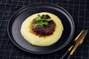 A medium rare ostrich steak in barbecue sauce on a cloud of herb mashed potato topped with fresh green arugula leaves on a black plate on a black checkered table cloth