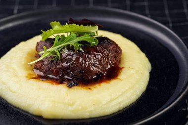 A medium rare ostrich steak in barbecue sauce on a cloud of herb mashed potato topped with fresh green arugula leaves on a black plate on a black checkered table cloth
