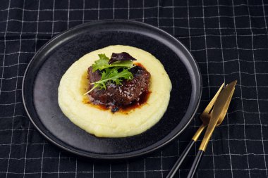 A medium rare ostrich steak in barbecue sauce on a cloud of herb mashed potato topped with fresh green arugula leaves on a black plate on a black checkered table cloth