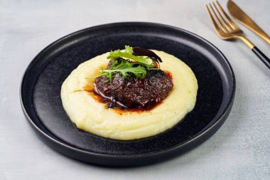 A medium rare ostrich steak in barbecue sauce on a cloud of herb mashed potato topped with fresh green arugula leaves on a black plate on a grey background