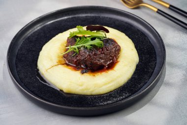 A medium rare ostrich steak in barbecue sauce on a cloud of herb mashed potato topped with fresh green arugula leaves on a black plate on a grey background