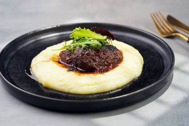 A medium rare ostrich steak in barbecue sauce on a cloud of herb mashed potato topped with fresh green arugula leaves on a black plate on a grey background