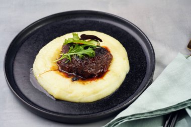 A medium rare ostrich steak in barbecue sauce on a cloud of herb mashed potato topped with fresh green arugula leaves on a black plate on a grey background