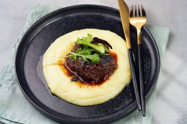 A medium rare ostrich steak in barbecue sauce on a cloud of herb mashed potato topped with fresh green arugula leaves on a black plate on a grey background