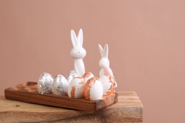 Easter decoration: eggs with silver and rose-gold metallic foil in a bamboos form on a wooden mediterranean style stool, white vase on beige colored seamless background, copy space