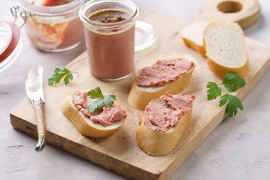 Homemade chicken liver pate on fresh french white wheat baguette slices on wooden board, glass mason jars with cooked liverwurst