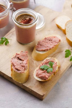 Homemade chicken liver pate on fresh french white wheat baguette slices on wooden board, glass mason jars with cooked liverwurst
