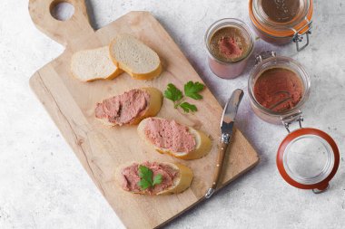 Homemade chicken liver pate on fresh french white wheat baguette slices on wooden plate, glass mason jars with cooked liverwurst, top view