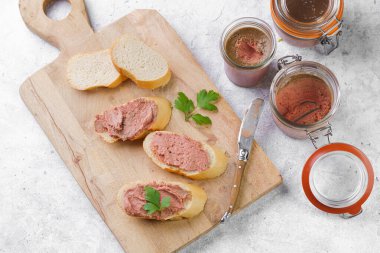 Homemade chicken liver pate on fresh french white wheat baguette slices on wooden plate, glass mason jars with cooked liverwurst, top view