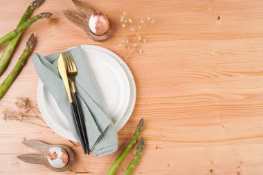 Boş beyaz seramik bir tabağın üst görüntüsü, keten peçetedeki altın çatal bıçak, tavşan şeklinde yumurta tutacağı, altın renkli yumurta tutacağı, ahşap arka planda taze kuşkonmaz, kopya alanı, üst manzara.