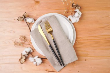Boş beyaz seramik bir tabağın üst görüntüsü, keten peçetedeki altın çatal bıçak takımı, ahşap arka plandaki pamuk çiçek süslemesi, fotokopi alanı, üst görünüm