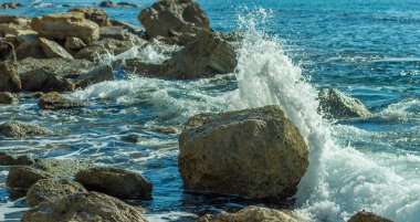 Bir Kıbrıs plajında kayalara çarpan deniz dalgalarının fotoğrafı, su sıçramaları, okyanus gelgitlerinin gücü ve güzelliğini yansıtıyor.