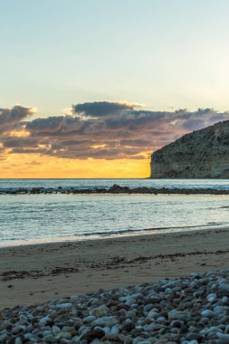 Bir Kıbrıs plajında gün batımının büyüleyici güzelliğine tanık olun, yumuşak pastel renklerle boyanmış, bulutlar denizin üzerinde süzülüyor ve heybetli kayalık bir uçurum, sakin ve resimli bir sahil manzarası yaratıyor.