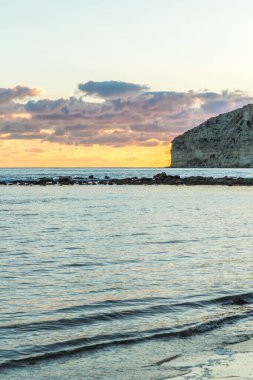 Bir Kıbrıs plajında gün batımının büyüleyici güzelliğine tanık olun, yumuşak pastel renklerle boyanmış, bulutlar denizin üzerinde süzülüyor ve heybetli kayalık bir uçurum, sakin ve resimli bir sahil manzarası yaratıyor.