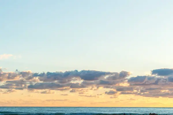 Bir Kıbrıs plajında gün batımının büyüleyici güzelliğine tanık olun, yumuşak pastel renklerle boyanmış, bulutlar denizin üzerinde süzülüyor ve heybetli kayalık bir uçurum, sakin ve resimli bir sahil manzarası yaratıyor.