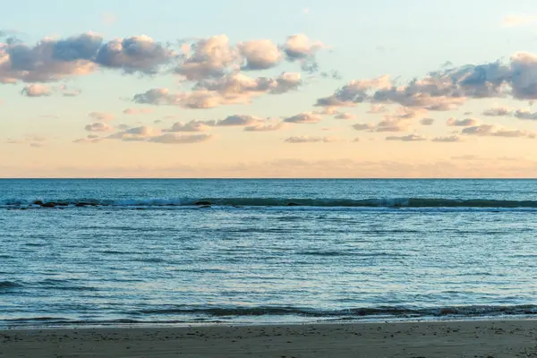 Bir Kıbrıs plajında gün batımının büyüleyici güzelliğine tanık olun, yumuşak pastel renklerle boyanmış, bulutlar denizin üzerinde süzülüyor ve heybetli kayalık bir uçurum, sakin ve resimli bir sahil manzarası yaratıyor.