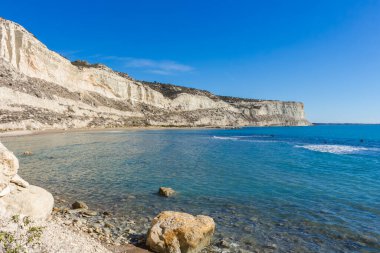 Arka planda kristal berraklığında mavi suları ve yüksek bir uçurumu olan kumlu bir sahil manzarası. Sakin su, beyaz kum ve kayalık ön plan dinlenmek ve güneşlenmek için ideal sakin bir kıyı manzarası yaratır.