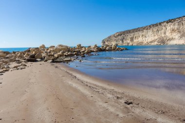 Arka planda kristal berraklığında mavi suları ve yüksek bir uçurumu olan kumlu bir sahil manzarası. Sakin su, beyaz kum ve kayalık ön plan dinlenmek ve güneşlenmek için ideal sakin bir kıyı manzarası yaratır.