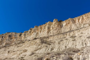 Arka planda kristal berraklığında mavi suları ve yüksek bir uçurumu olan kumlu bir sahil manzarası. Sakin su, beyaz kum ve kayalık ön plan dinlenmek ve güneşlenmek için ideal sakin bir kıyı manzarası yaratır.