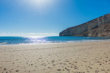 Arka planda kristal berraklığında mavi suları ve yüksek bir uçurumu olan kumlu bir sahil manzarası. Sakin su, beyaz kum ve kayalık ön plan dinlenmek ve güneşlenmek için ideal sakin bir kıyı manzarası yaratır.