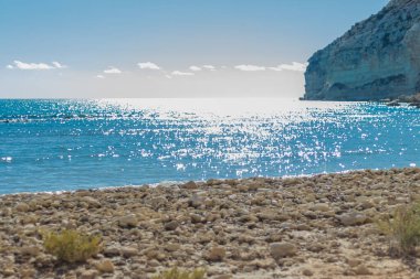 Arka planda kristal berraklığında mavi suları ve yüksek bir uçurumu olan kumlu bir sahil manzarası. Sakin su, beyaz kum ve kayalık ön plan dinlenmek ve güneşlenmek için ideal sakin bir kıyı manzarası yaratır.