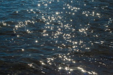 Güneşli suyun dingin güzelliğini yansıtan bir görüntü. Güneş ışığının dalgalanan yüzeyindeki yansımalarıyla oluşan ışıltılı bir efekt.
