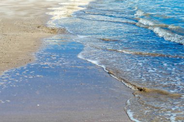 Görüntü, yumuşak dalgaların kumlu bir kıyı şeridini okşayarak berrak mavi suya doğru çaprazlama uzandığı sakin bir kıyı sahnesi yakalıyor. Islak kumda yansımalar derinlik ve görsel ilgi ekler.
