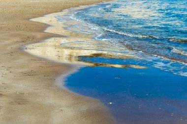 Görüntü, yumuşak dalgaların kumlu bir kıyı şeridini okşayarak berrak mavi suya doğru çaprazlama uzandığı sakin bir kıyı sahnesi yakalıyor. Islak kumda yansımalar derinlik ve görsel ilgi ekler.