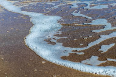 Görüntü, yumuşak dalgaların kumlu bir kıyı şeridini okşayarak berrak mavi suya doğru çaprazlama uzandığı sakin bir kıyı sahnesi yakalıyor. Islak kumda yansımalar derinlik ve görsel ilgi ekler.