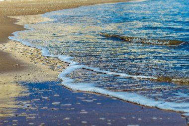 Görüntü, yumuşak dalgaların kumlu bir kıyı şeridini okşayarak berrak mavi suya doğru çaprazlama uzandığı sakin bir kıyı sahnesi yakalıyor. Islak kumda yansımalar derinlik ve görsel ilgi ekler.