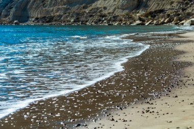 Kıbrıs 'ta sakin ve büyüleyici bir plaj sahnesi, berrak mavi gökyüzü, turkuaz suların güneş ışığında ışıldaması, sıcak kahverengi kumlar ve kıyıya vuran nazik dalgaların yer aldığı. Fotoğraf huzur ve sükuneti simgeliyor.