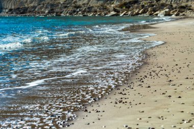 Kıbrıs 'ta sakin ve büyüleyici bir plaj sahnesi, berrak mavi gökyüzü, turkuaz suların güneş ışığında ışıldaması, sıcak kahverengi kumlar ve kıyıya vuran nazik dalgaların yer aldığı. Fotoğraf huzur ve sükuneti simgeliyor.