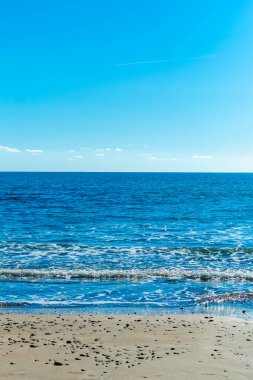 Kıbrıs 'ta sakin ve büyüleyici bir plaj sahnesi, berrak mavi gökyüzü, turkuaz suların güneş ışığında ışıldaması, sıcak kahverengi kumlar ve kıyıya vuran nazik dalgaların yer aldığı. Fotoğraf huzur ve sükuneti simgeliyor.