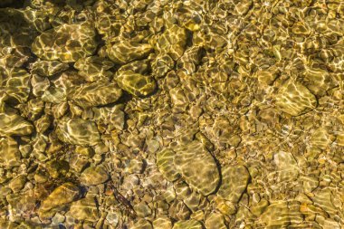 Sığ bir su kütlesinin dibini, muhtemelen bir akarsu ya da nehri, güneş ışığının filtreleyerek temiz yüzey üzerinde dalgalanma etkisi oluşturduğu sakin bir bakış. Alttaki kayaların şekli bozulmuş.