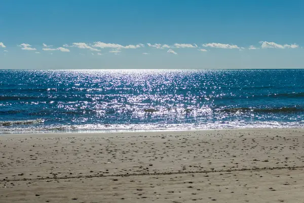 Kıbrıs 'ta sakin ve büyüleyici bir plaj sahnesi, berrak mavi gökyüzü, turkuaz suların güneş ışığında ışıldaması, sıcak kahverengi kumlar ve kıyıya vuran nazik dalgaların yer aldığı. Fotoğraf huzur ve sükuneti simgeliyor.